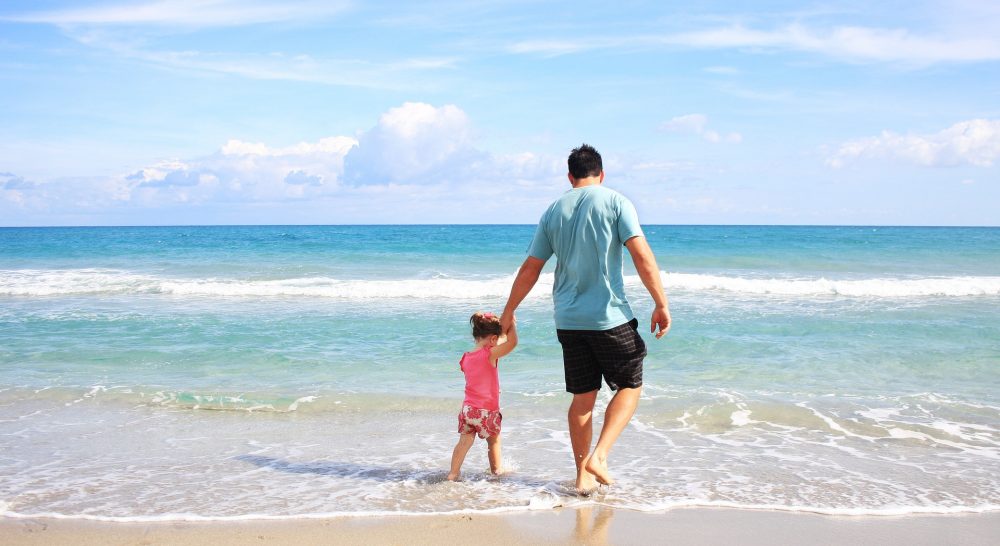 Man holding hand of child whilst they walk across the shore of the beach