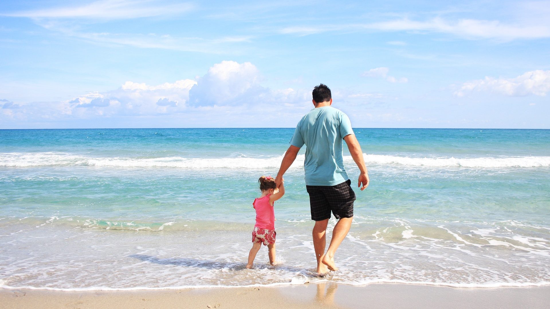 Man holding hand of child whilst they walk across the shore of the beach